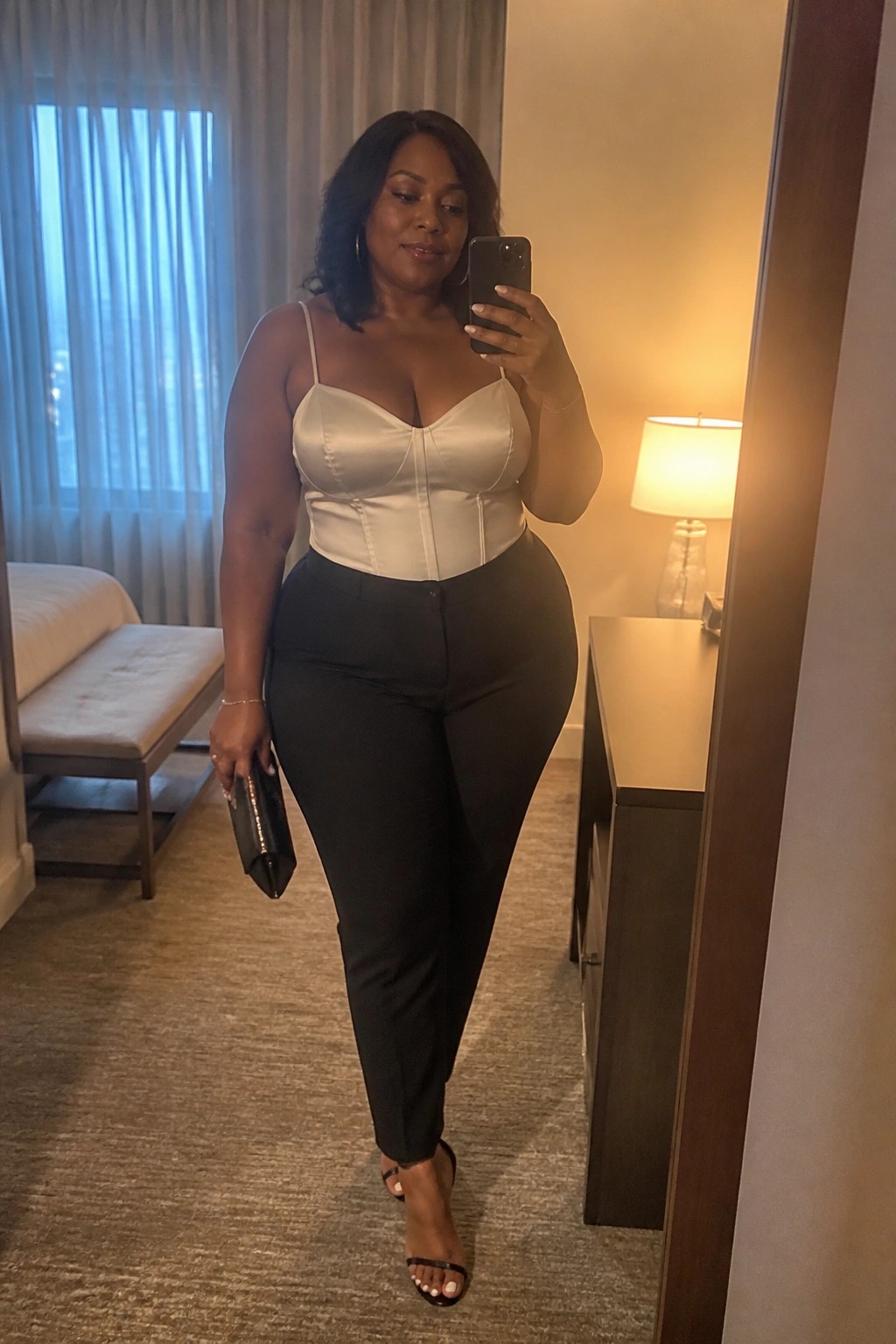 Woman in white satin corset top and black pants taking mirror selfie in hotel room