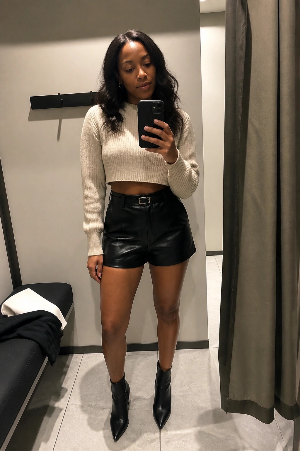 Woman in cream cropped sweater and black leather shorts taking mirror selfie in fitting room