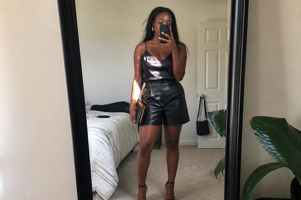 Woman in satin camisole and black leather shorts taking bedroom mirror selfie