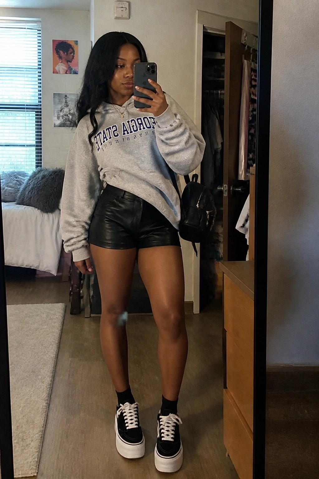 Woman in gray Georgia State hoodie and black leather shorts taking mirror selfie in bedroom