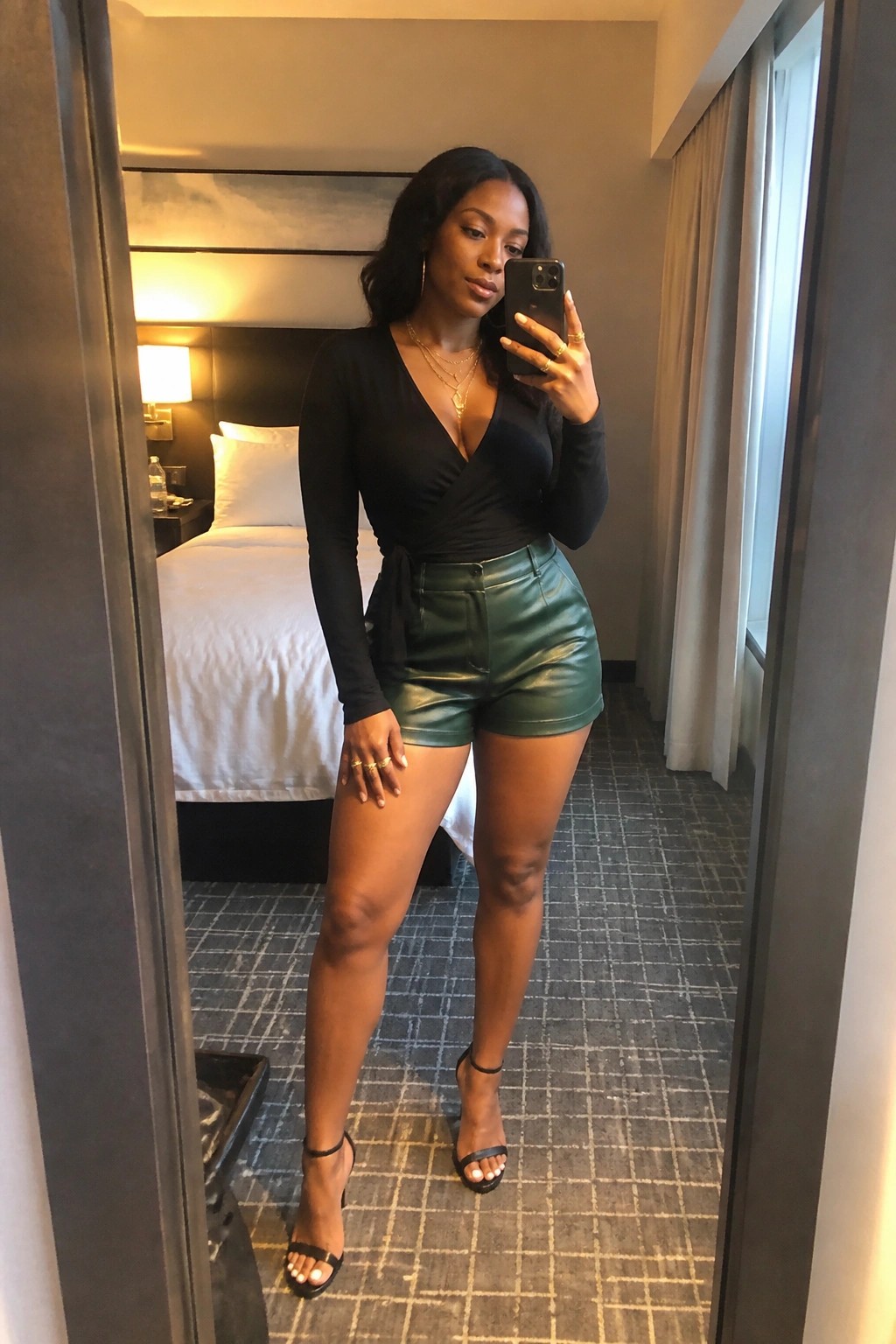 Woman in black wrap top and green leather shorts taking mirror selfie in hotel room.