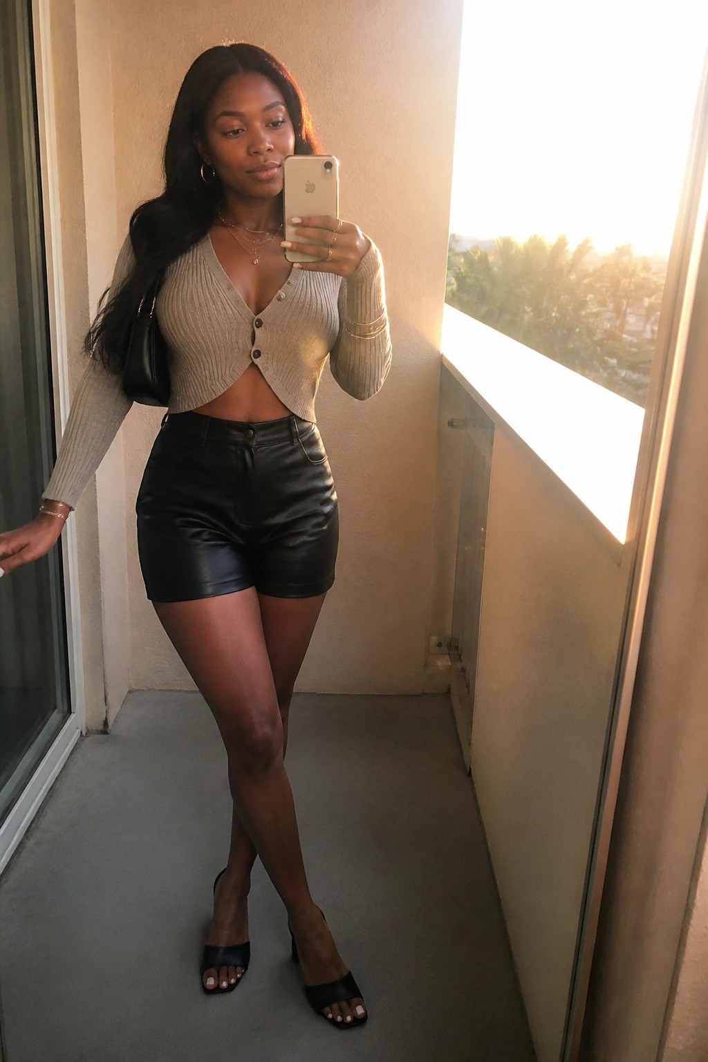 Woman in beige cropped cardigan and black leather shorts taking balcony selfie at sunset.