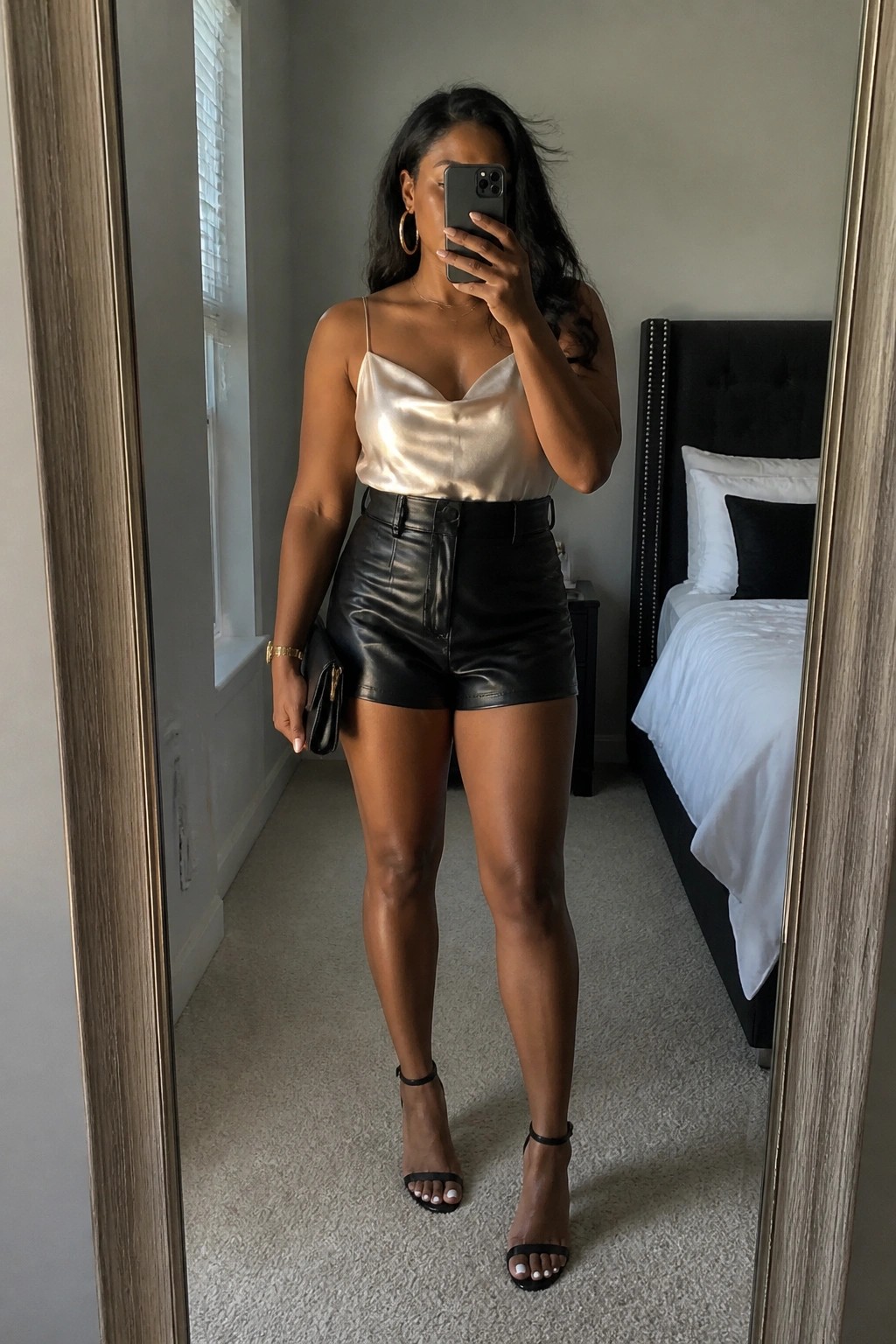 Woman in satin camisole and black leather shorts taking bedroom mirror selfie