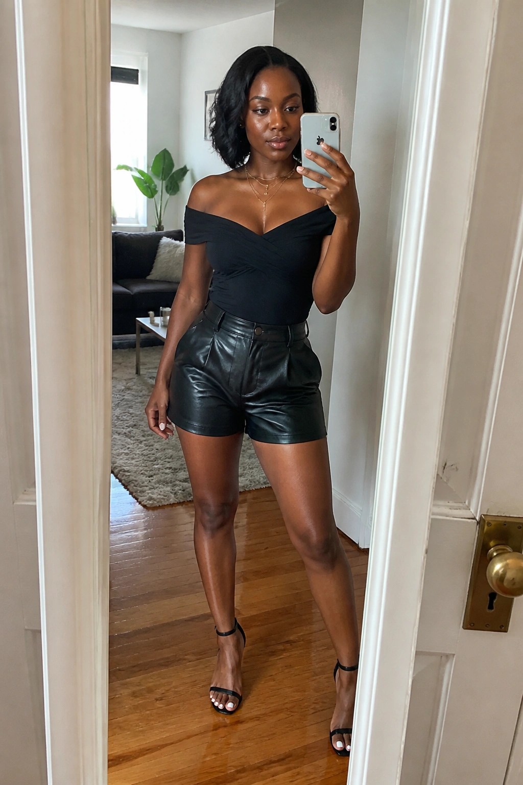 Woman in black off-shoulder top and leather shorts taking mirror selfie with phone