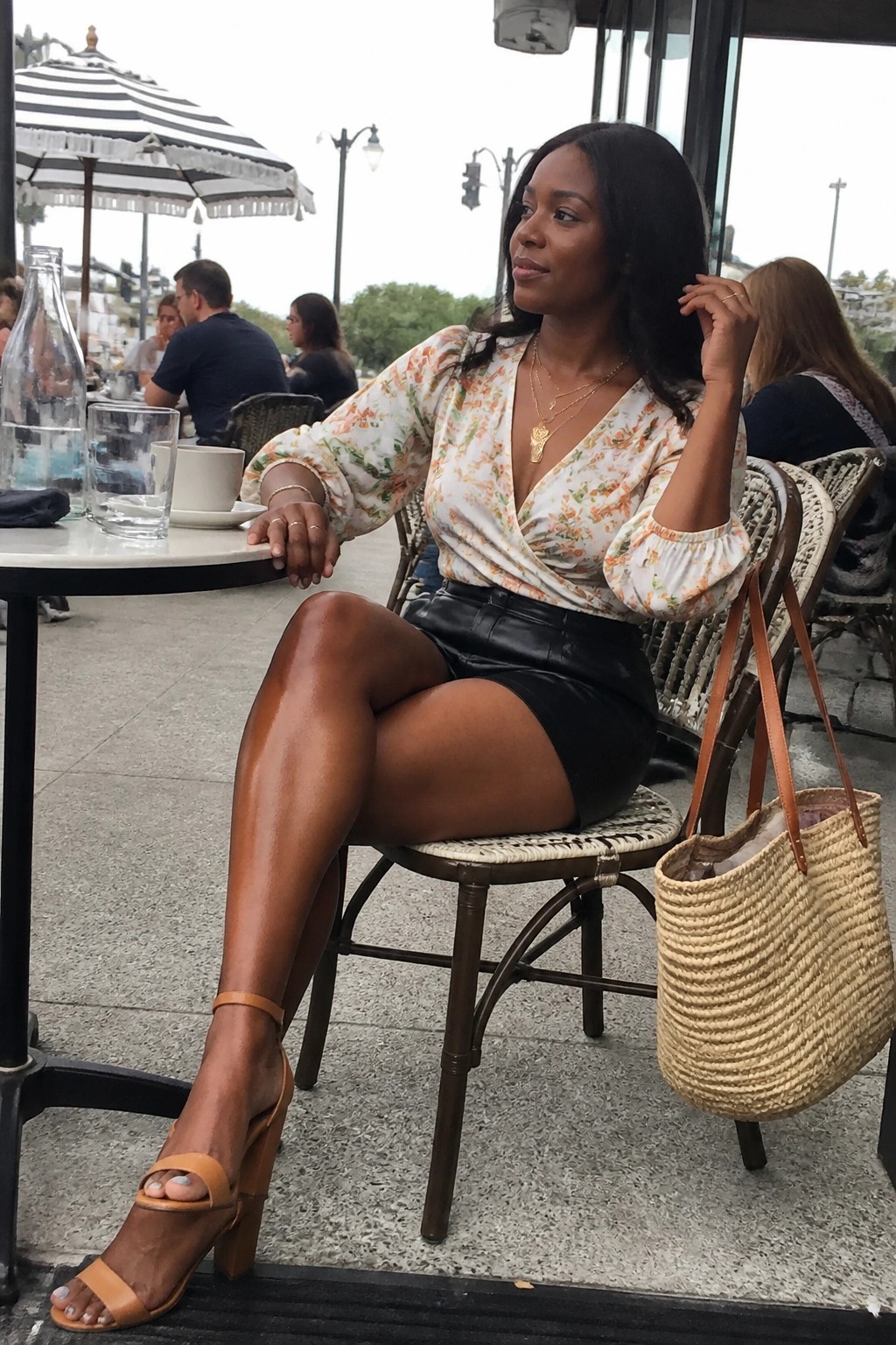 Woman in floral wrap top and black leather skirt at outdoor cafe with straw bag