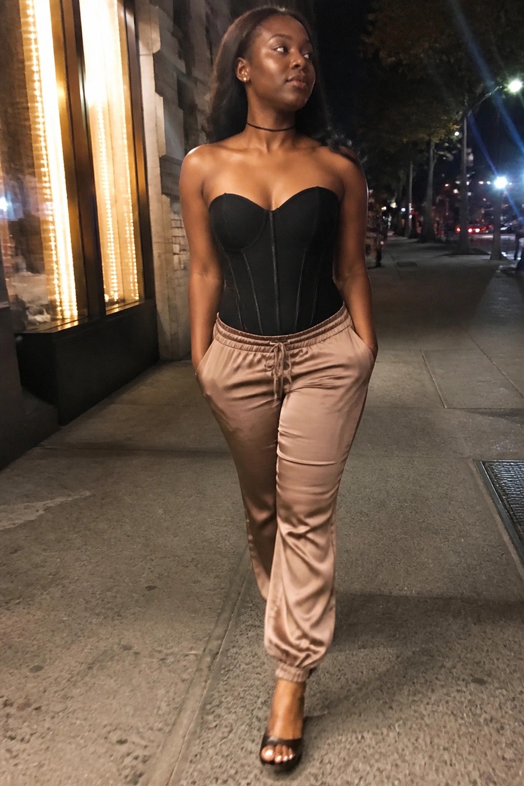 Woman in black strapless corset and beige satin joggers on nighttime city sidewalk.