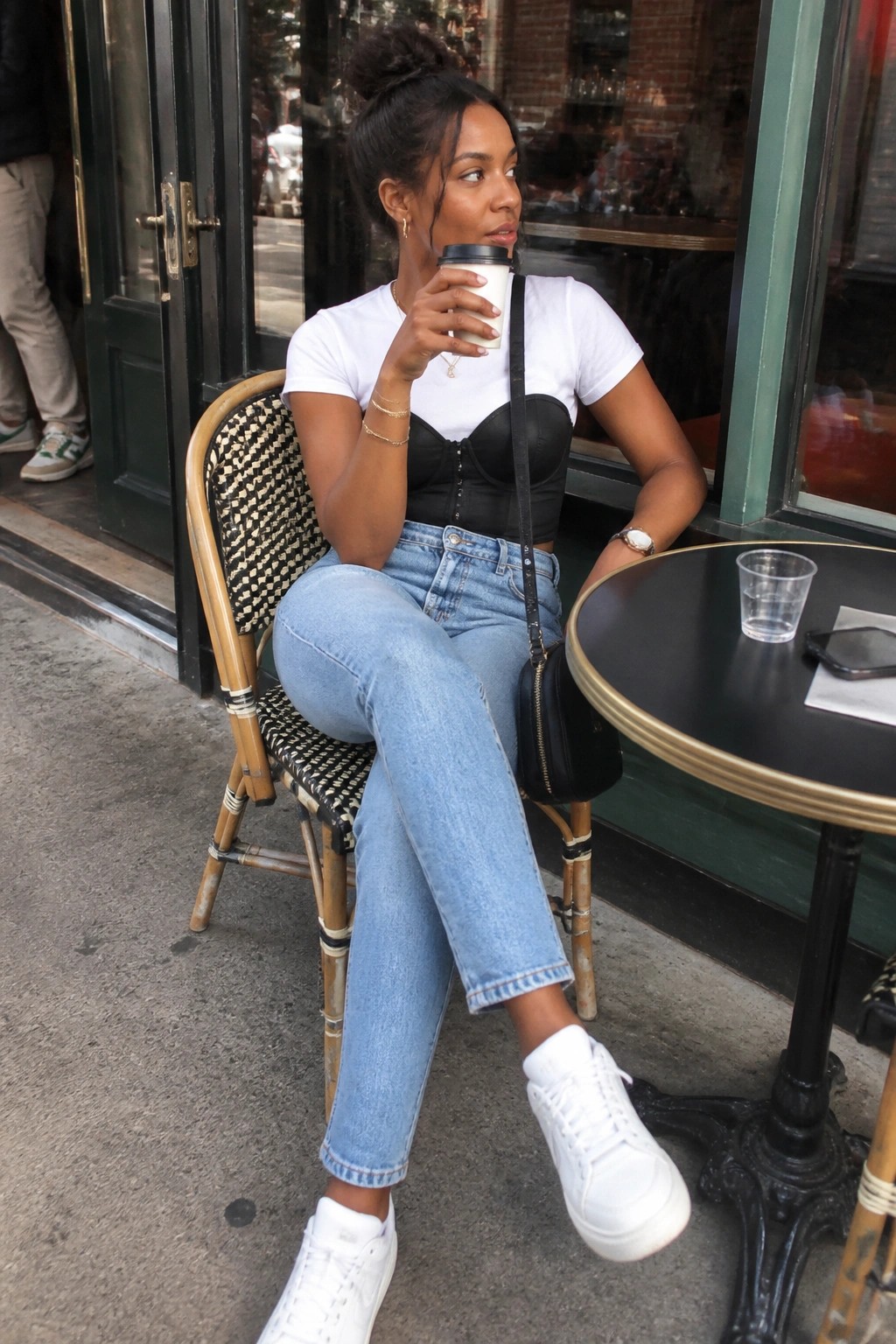 Woman in white tee over black corset sips coffee at sidewalk cafe.