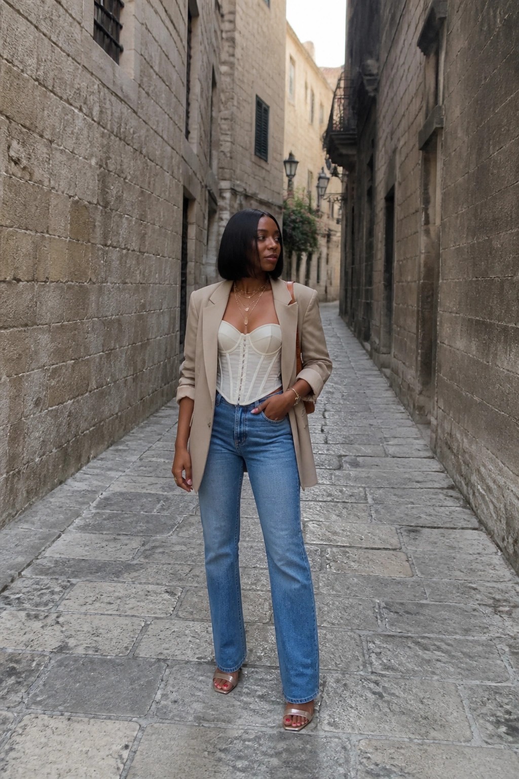 Woman in beige blazer over white corset and blue jeans in stone alley.