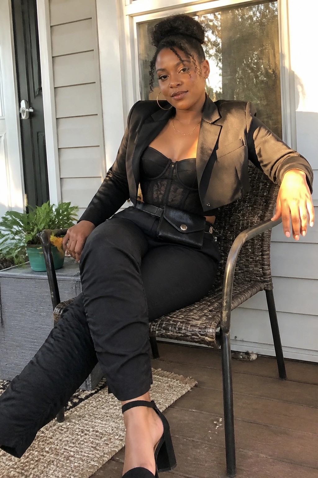 Black woman in satin blazer and lace corset seated on wicker porch chair