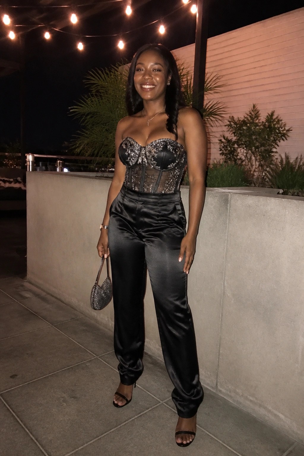 Woman in black sequined corset top and satin pants holding beaded purse at night.