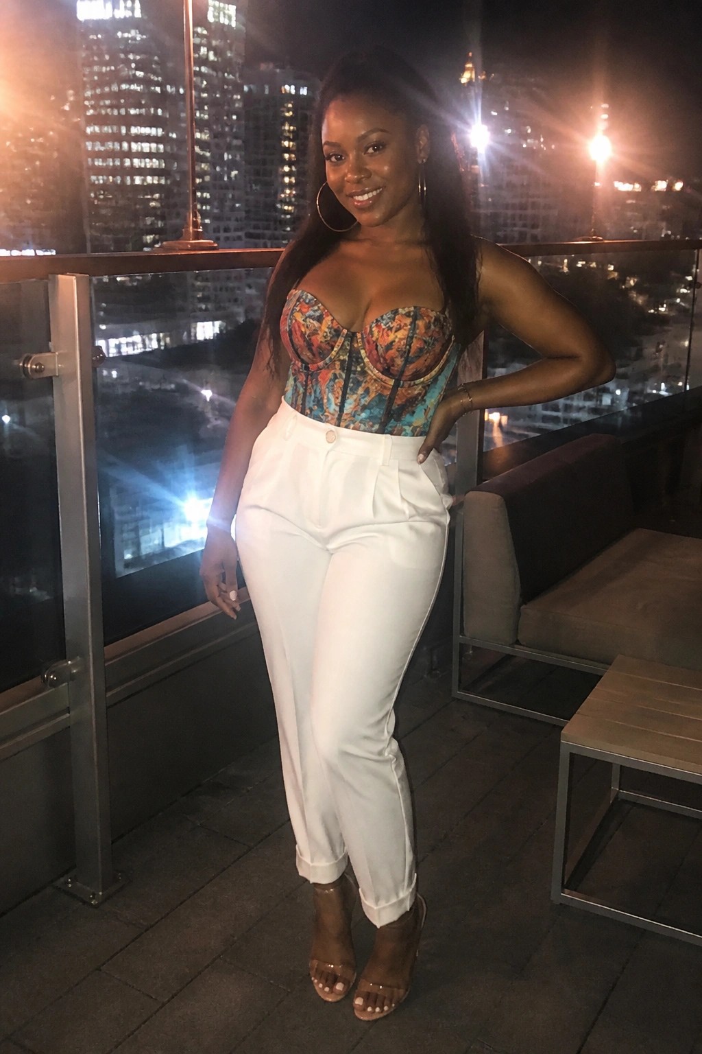 Woman in vibrant corset top and white pants posing on nighttime city balcony