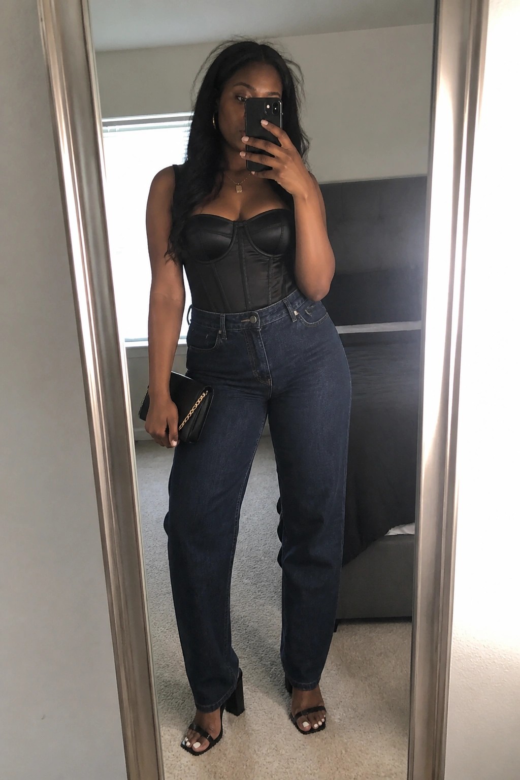 Woman wearing a black strapless corset top tucked into dark blue wide leg jeans with black block heel sandals and holding a small black clutch while taking a mirror selfie.