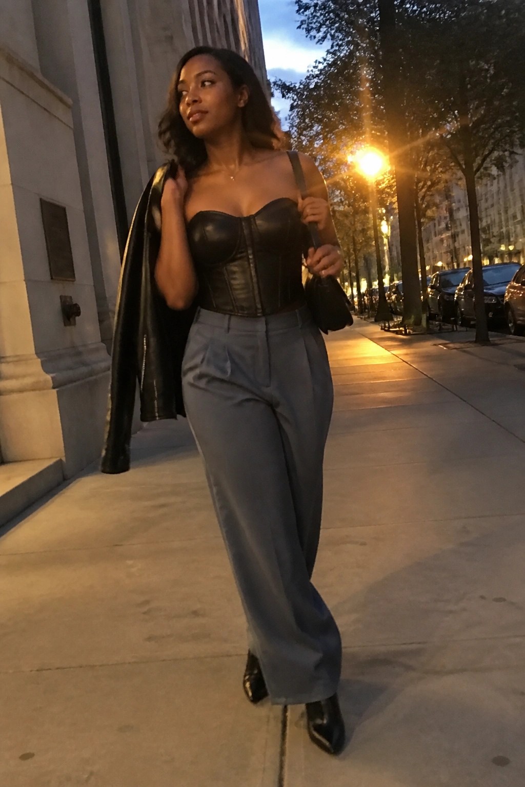 Woman in black corset and gray trousers holding jacket on city sidewalk at night
