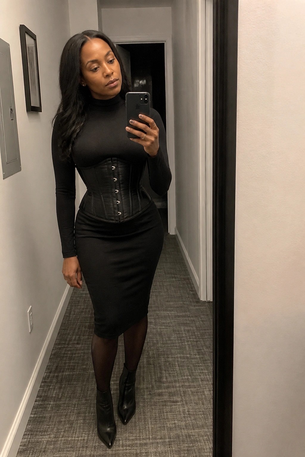 Woman in black corset, turtleneck, pencil skirt, and boots taking mirror selfie with iPhone