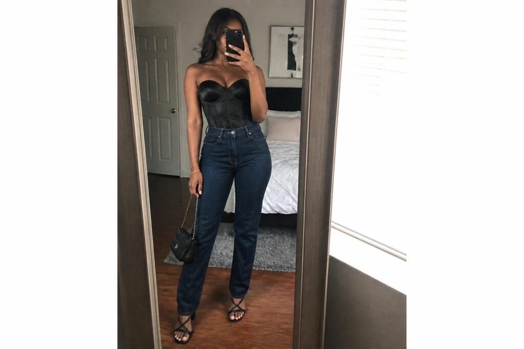 25 Fierce Girls’ Night Out Corset Outfit Ideas for Black Women Woman wearing a black strapless corset top tucked into dark blue wide leg jeans with black block heel sandals and holding a small black clutch while taking a mirror selfie.