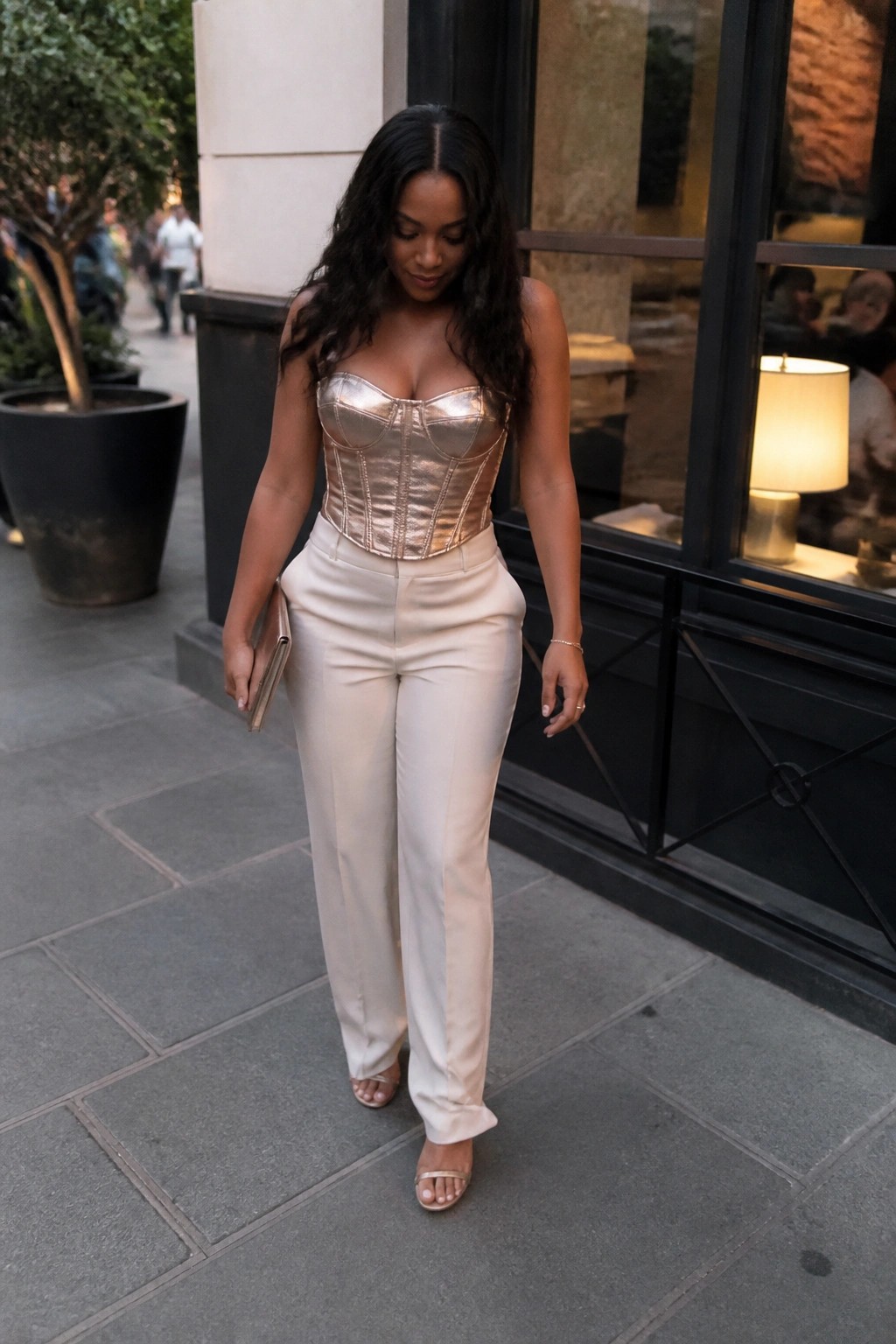 Woman in metallic gold corset and white pants walking on urban sidewalk
