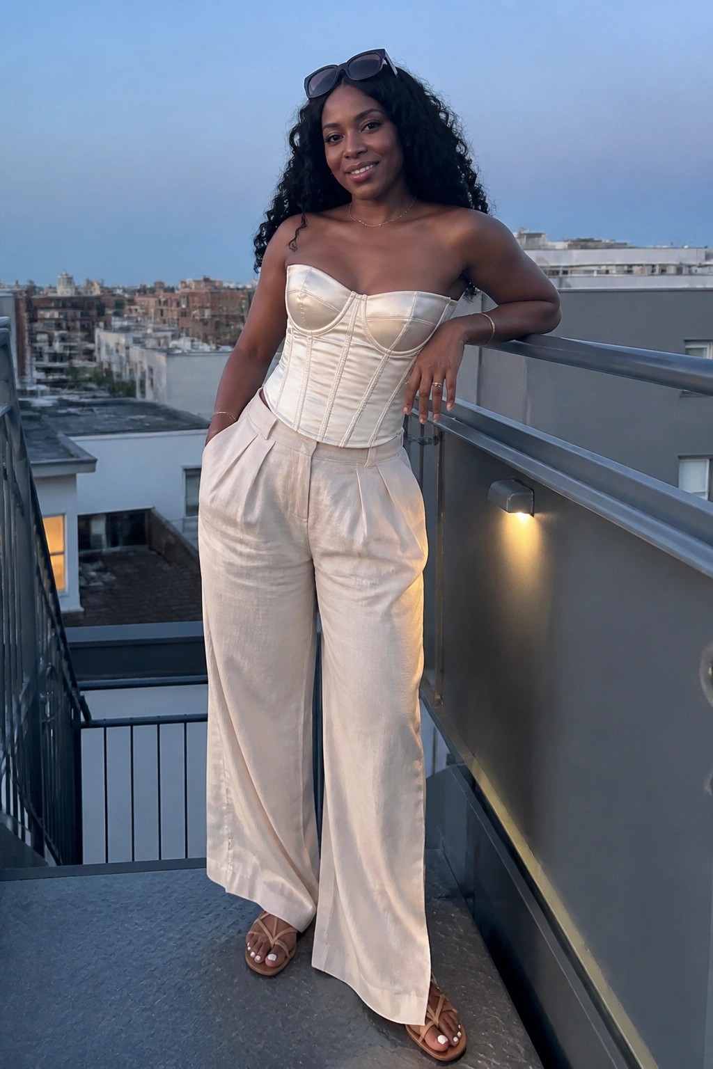 Woman in white strapless corset and wide-leg pants on city balcony at dusk