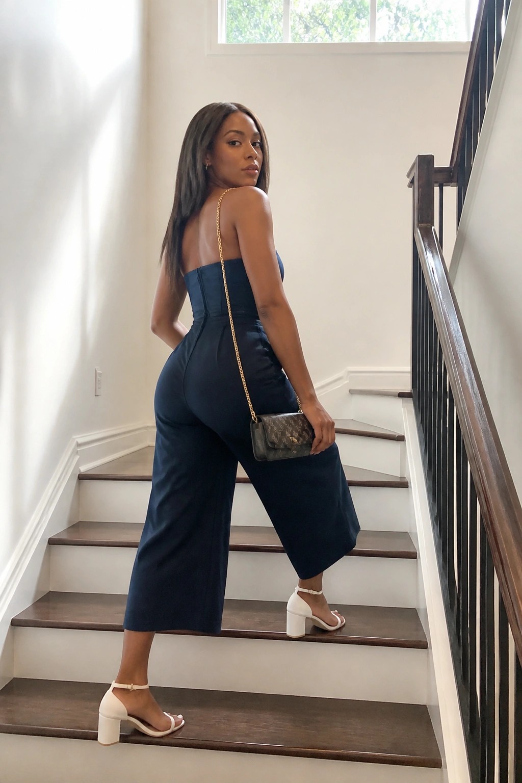 Woman in navy strapless jumpsuit with handbag on staircase in white heels
