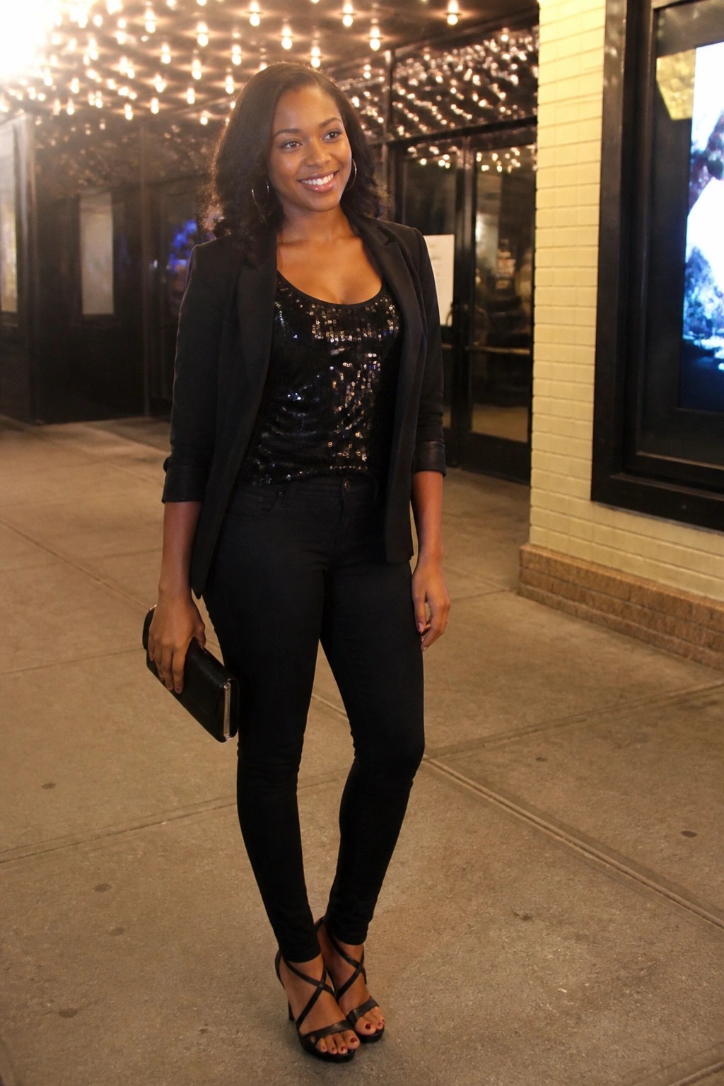 Smiling woman in black sequined top, blazer, and jeans holds clutch at night.