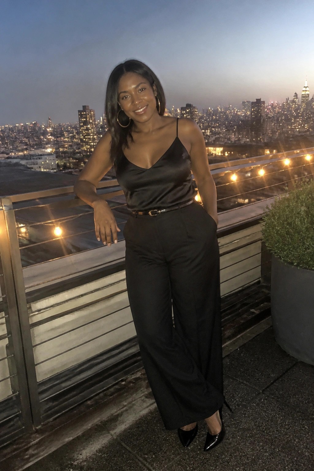 Woman in black satin top and wide-leg pants on rooftop overlooking city skyline at dusk
