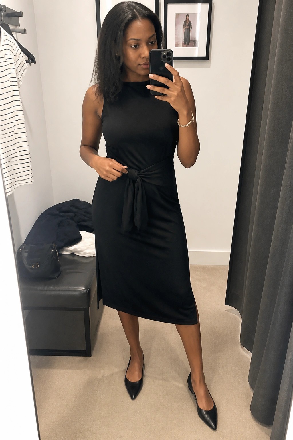 Woman in black sleeveless midi dress with waist tie taking mirror selfie in fitting room