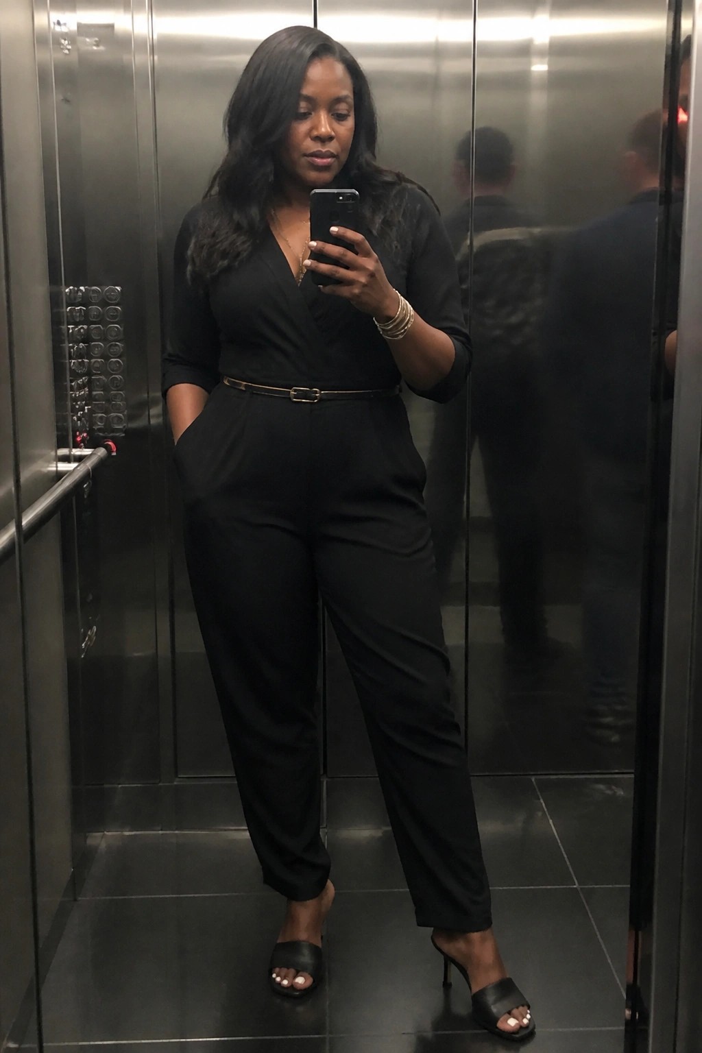 Woman in black jumpsuit taking mirror selfie with phone in elevator