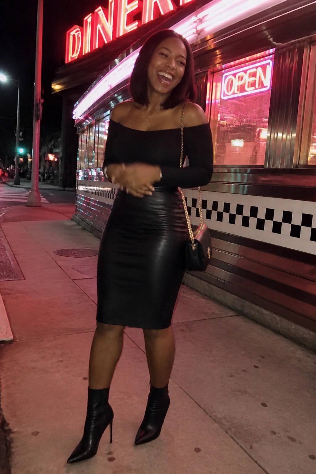 Smiling woman in black off-shoulder top and leather skirt outside neon diner at night.