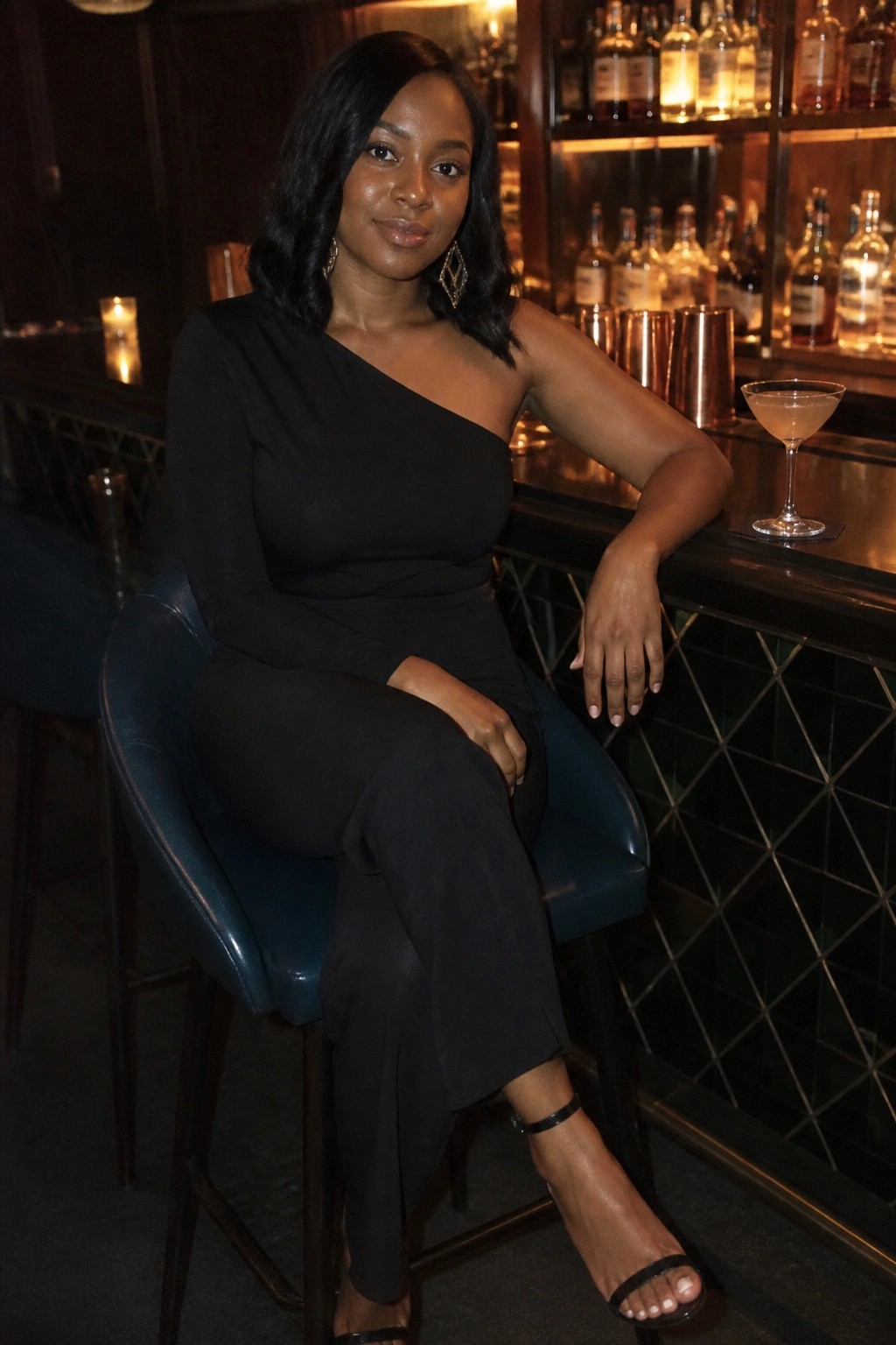 Woman in black one-shoulder jumpsuit seated at blue barstool with cocktail