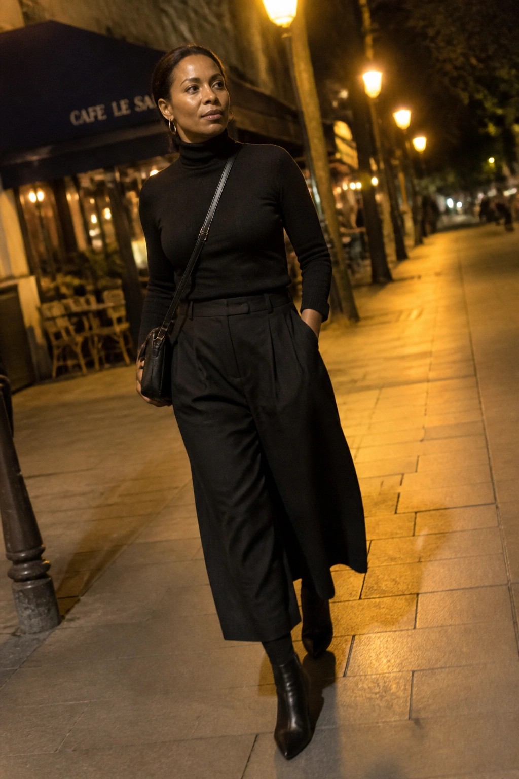 Woman in black turtleneck and wide pants with crossbody bag on lit sidewalk.