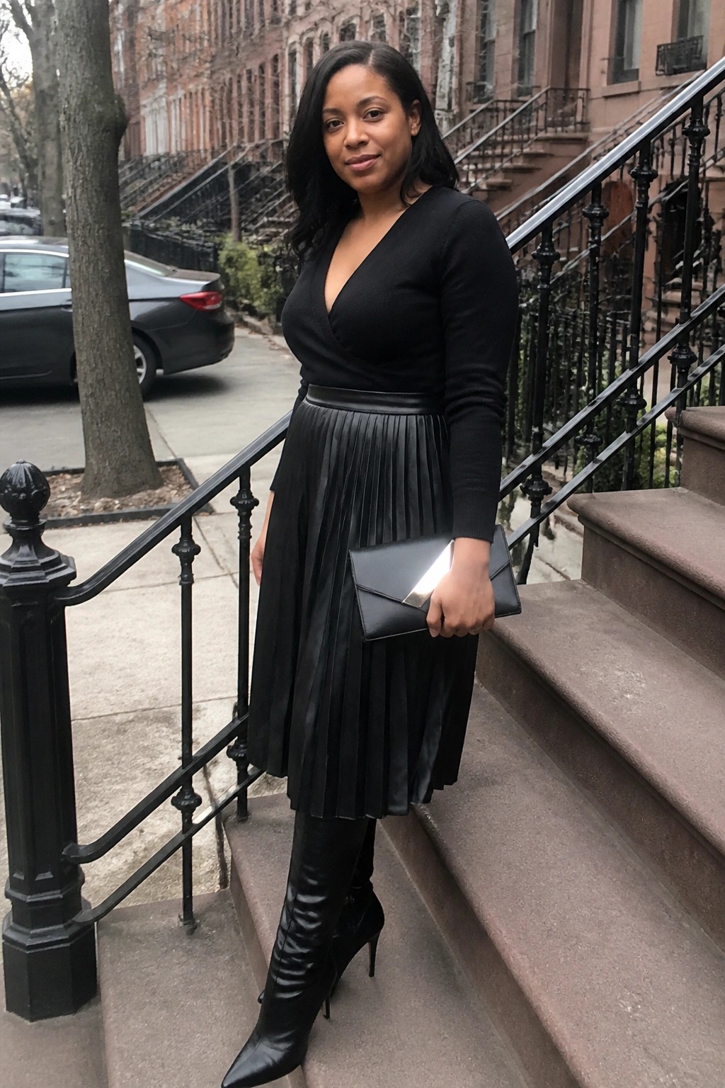 Woman wearing a black wrap V-neck sweater tucked into a black pleated faux-leather midi skirt with a wide belt, paired with black pointed-toe knee-high boots and holding a black envelope clutch while standing on outdoor steps.
