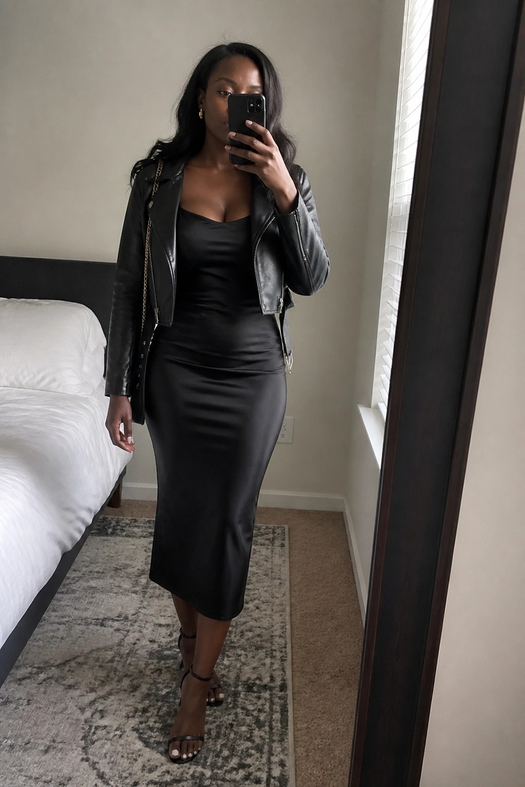 Woman in black leather jacket and satin midi dress taking bedroom mirror selfie