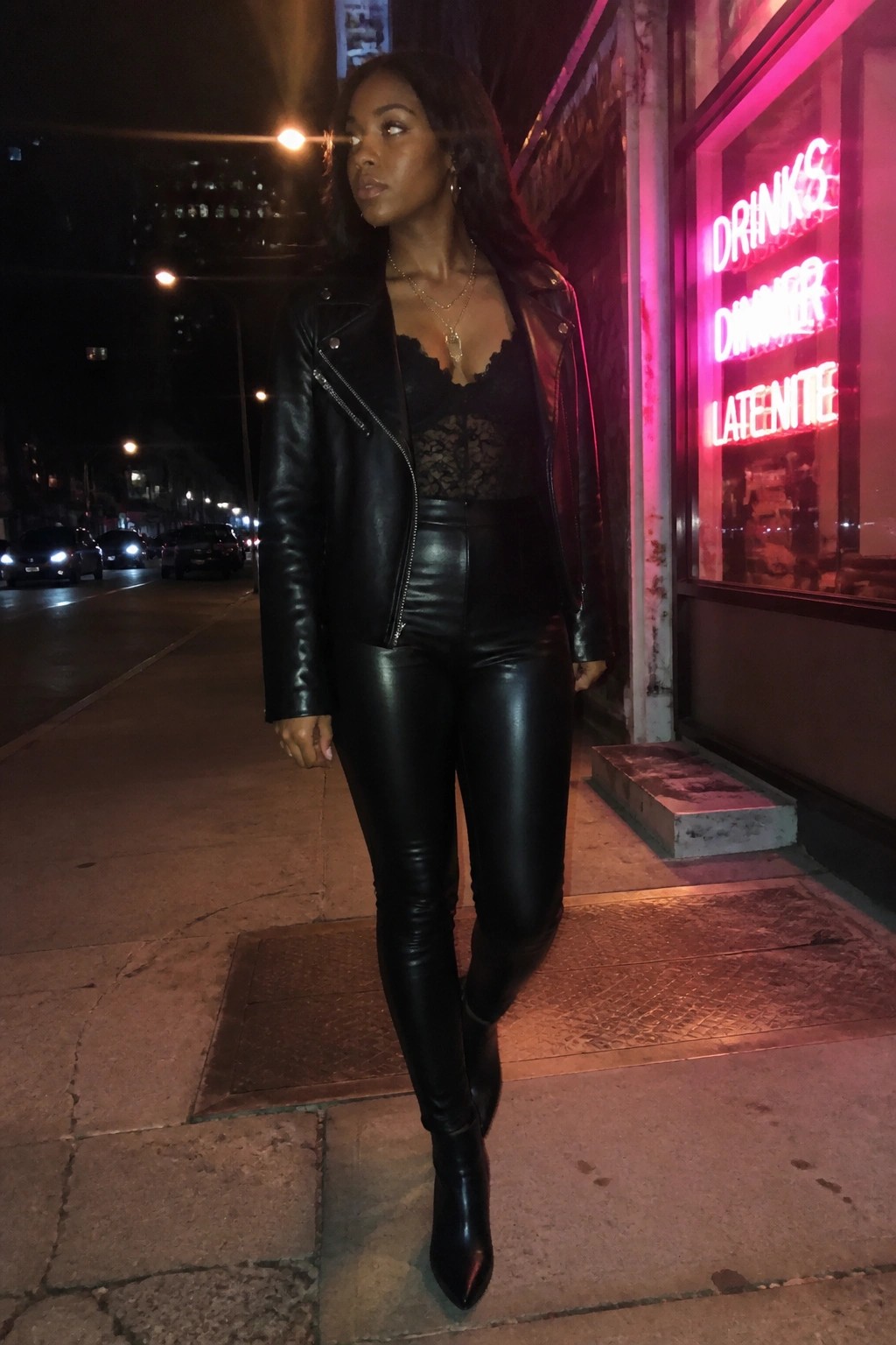 Woman in black leather jacket and shiny pants on neon-lit city sidewalk at night.