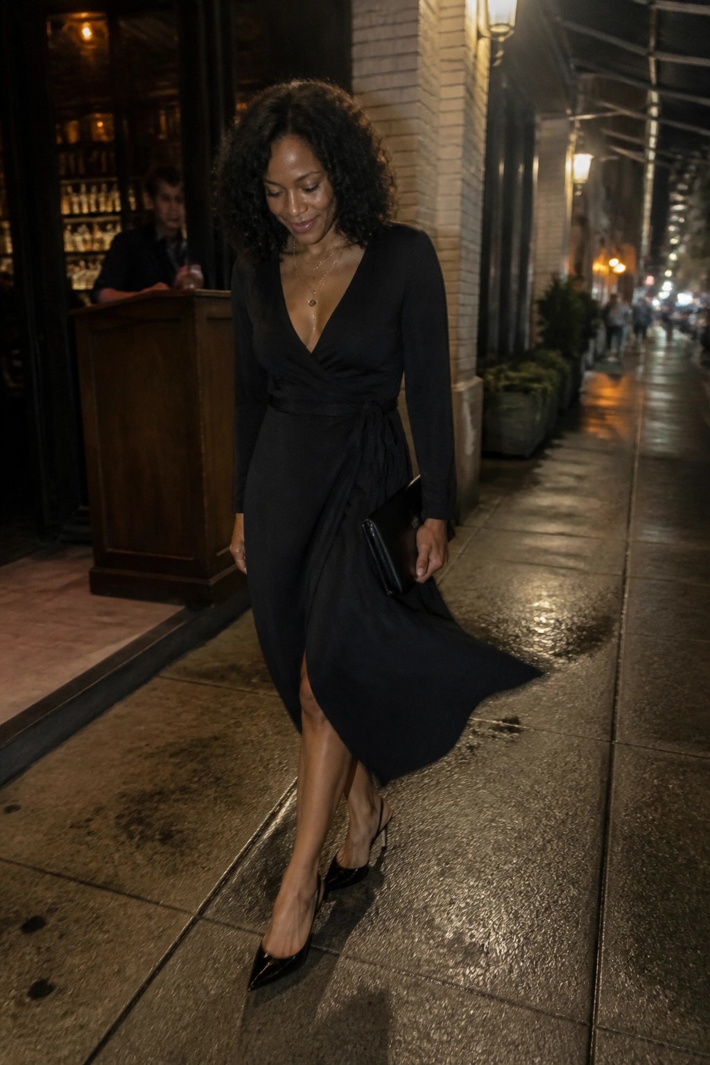 Woman in black wrap dress and heels on wet nighttime sidewalk