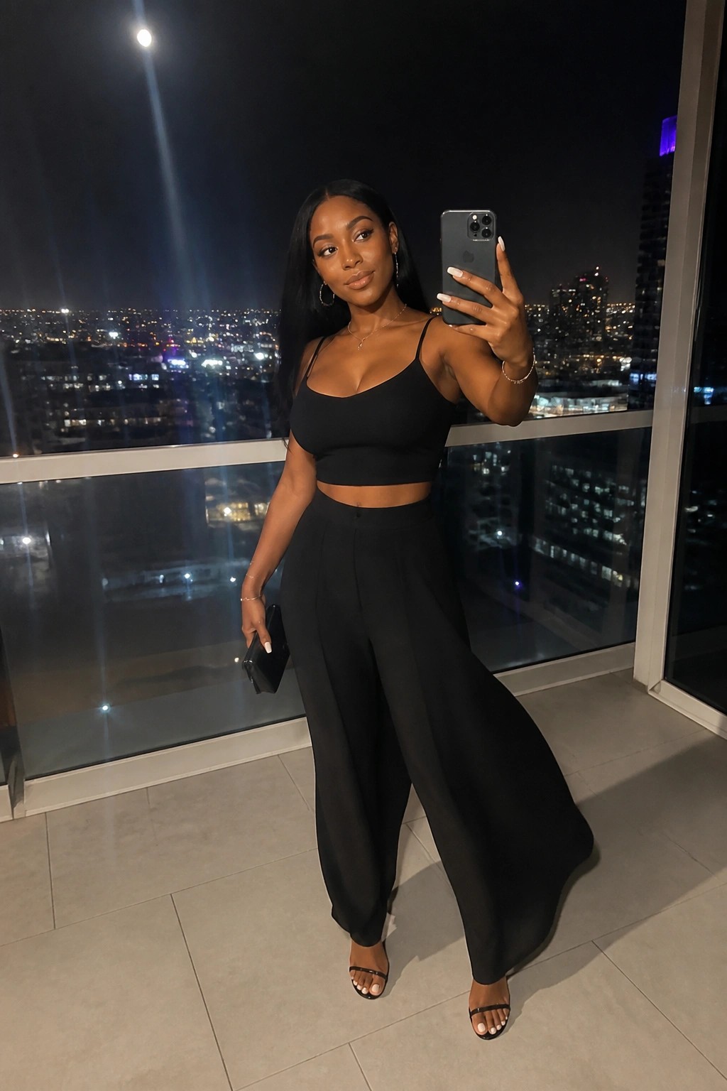 Woman in black crop top and wide-leg pants taking balcony selfie at night