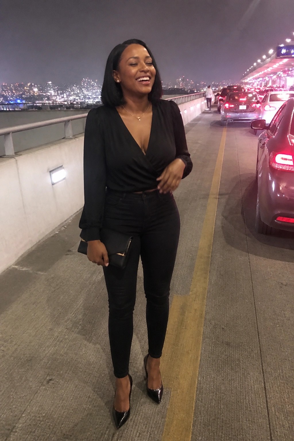 Smiling woman in black wrap top and jeans on nighttime city bridge with traffic.