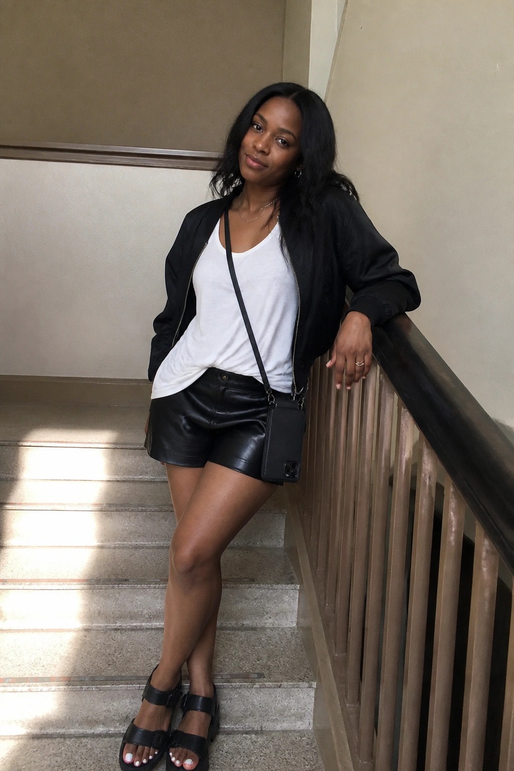 Woman in black leather shorts, white tank top, open black bomber jacket, black crossbody bag, and black sandals posing on indoor stairs.