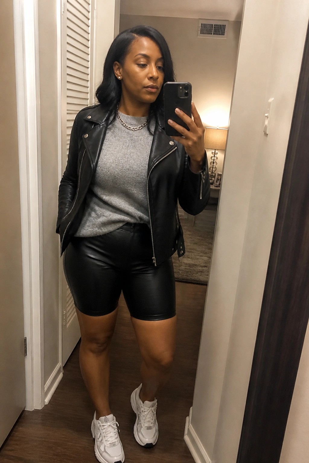 Woman wearing black leather moto jacket over gray sweater with black leather shorts, white sneakers, and silver chain necklace in mirror selfie.