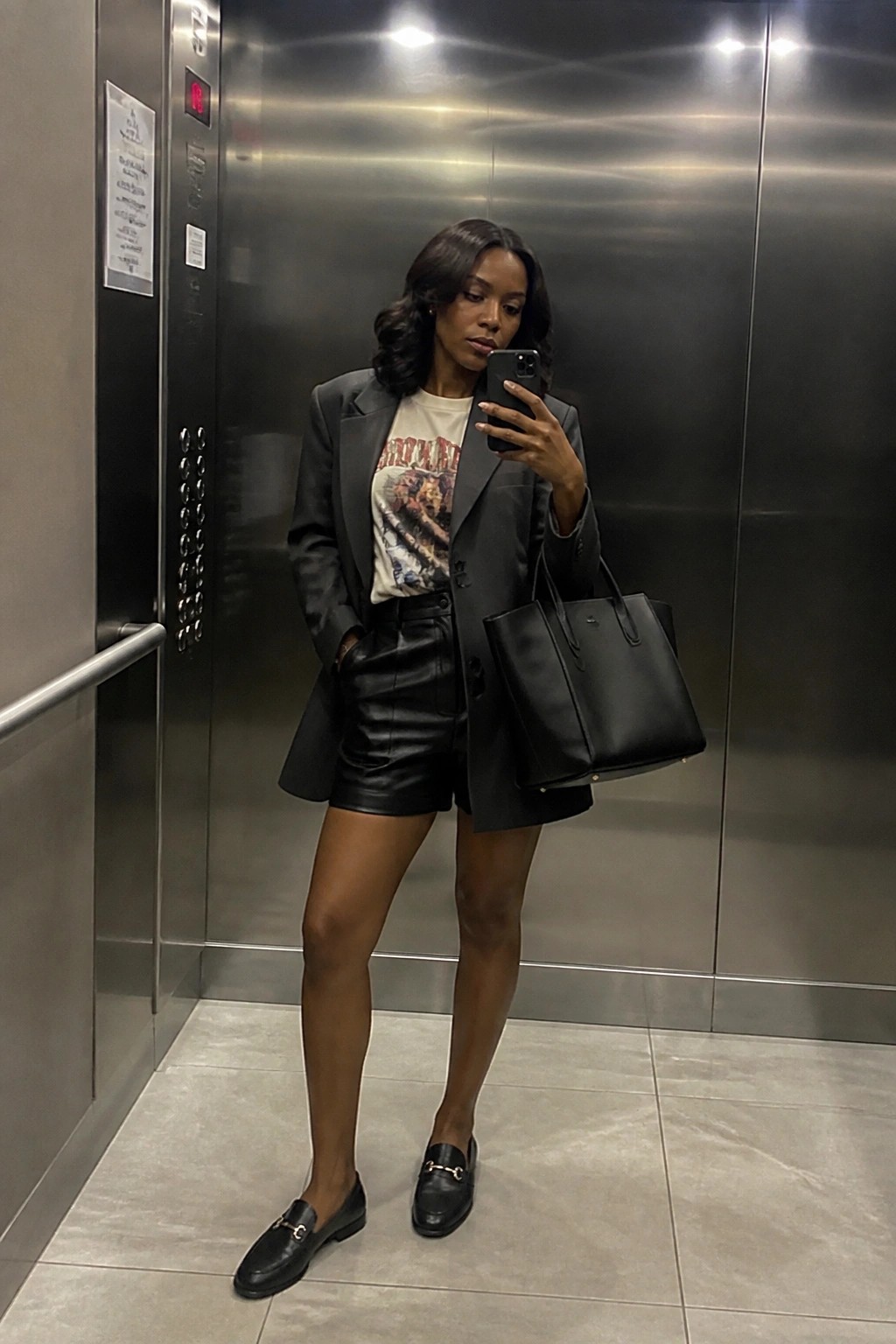 Woman in black blazer, graphic tee, and leather shorts taking selfie in elevator with bag