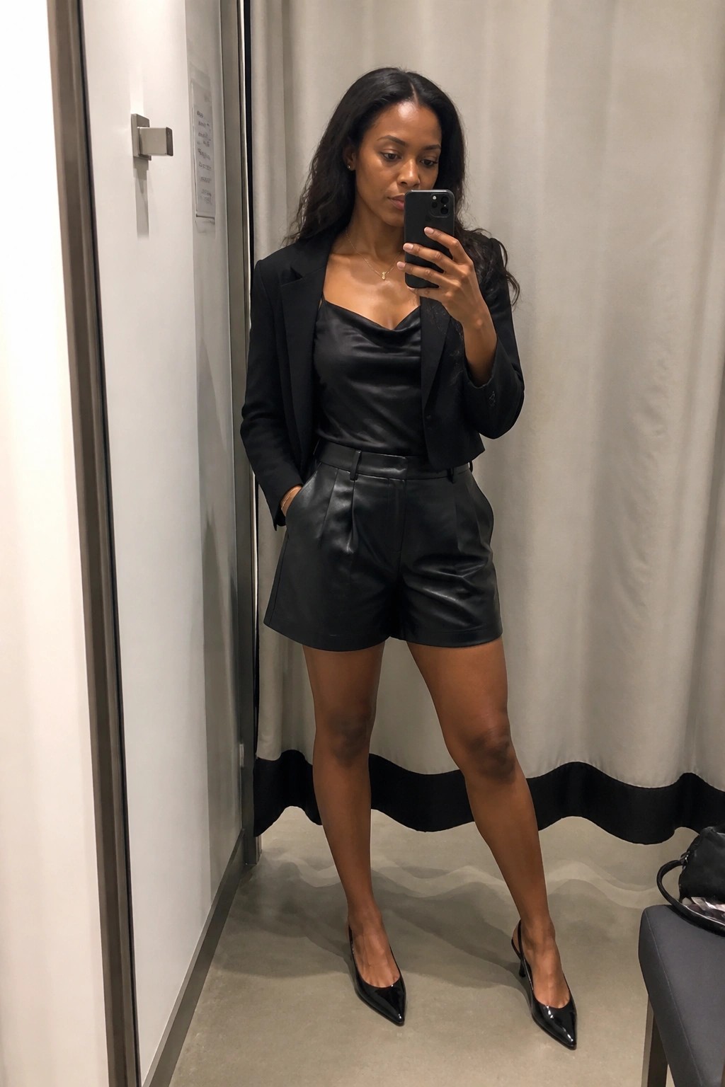 Woman in black blazer, satin top, and leather shorts taking mirror selfie in fitting room.