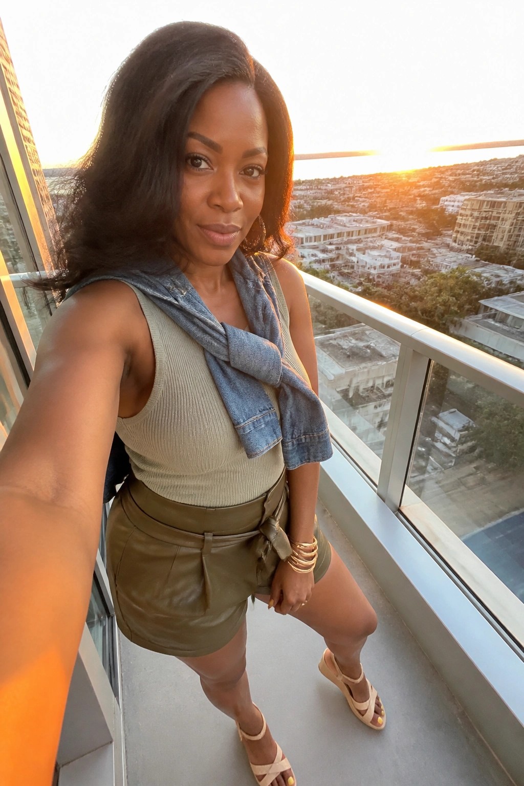 Woman in beige top and green shorts with denim jacket on balcony at sunset
