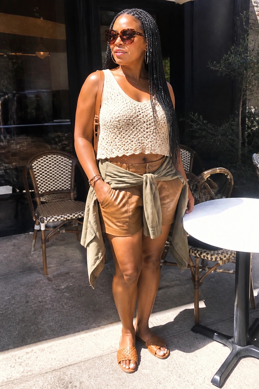 Woman in beige crochet crop top, brown leather shorts, green jacket tied at waist outdoors